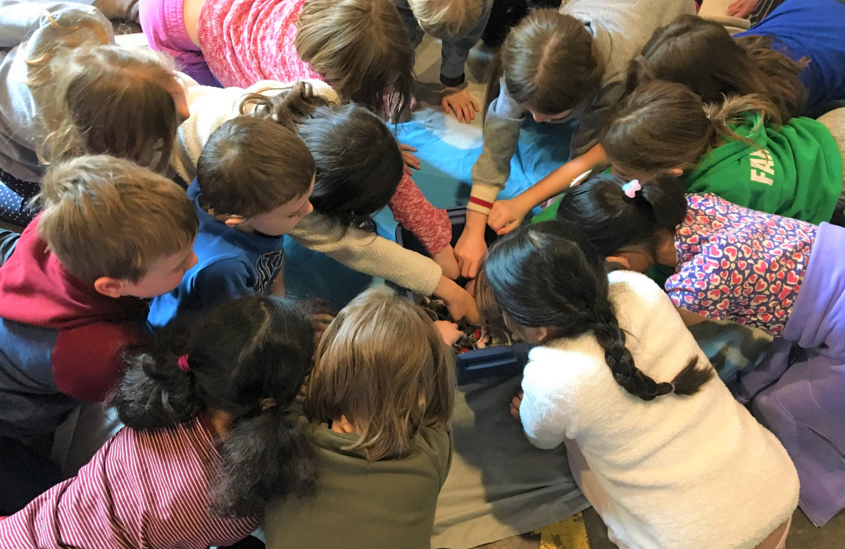 Children are in a circle leaning over a bin. Many of the children have their hands in the bin.